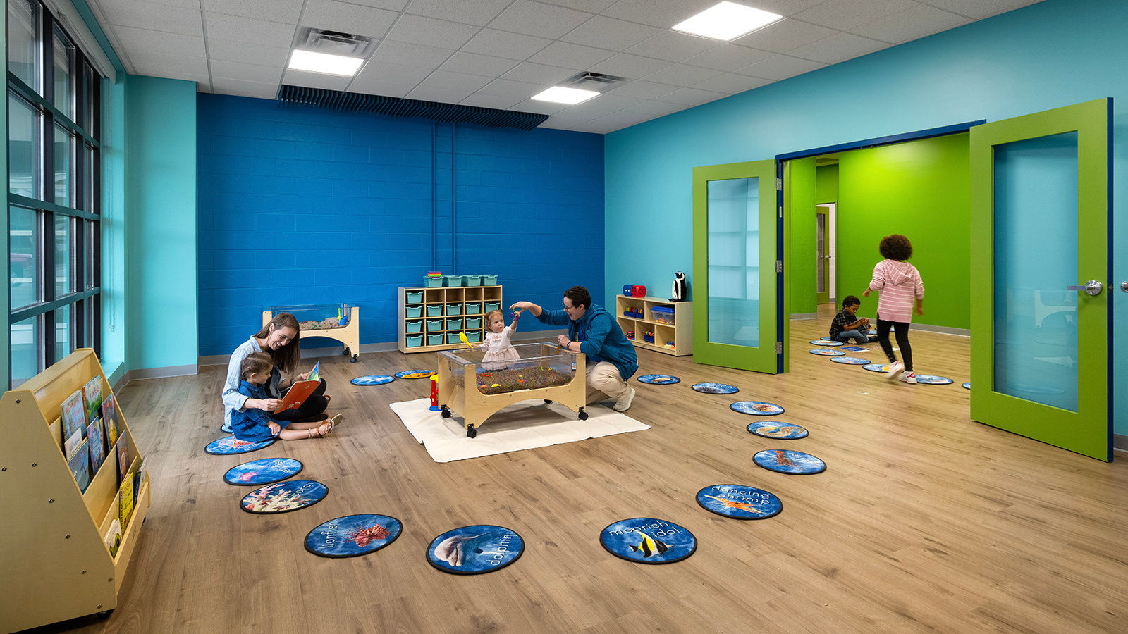 A classroom for WAVE Foundation featuring a mom and child reading a book, an employee playing with a young girl at the water table and two kids playing in the green classroom next door.