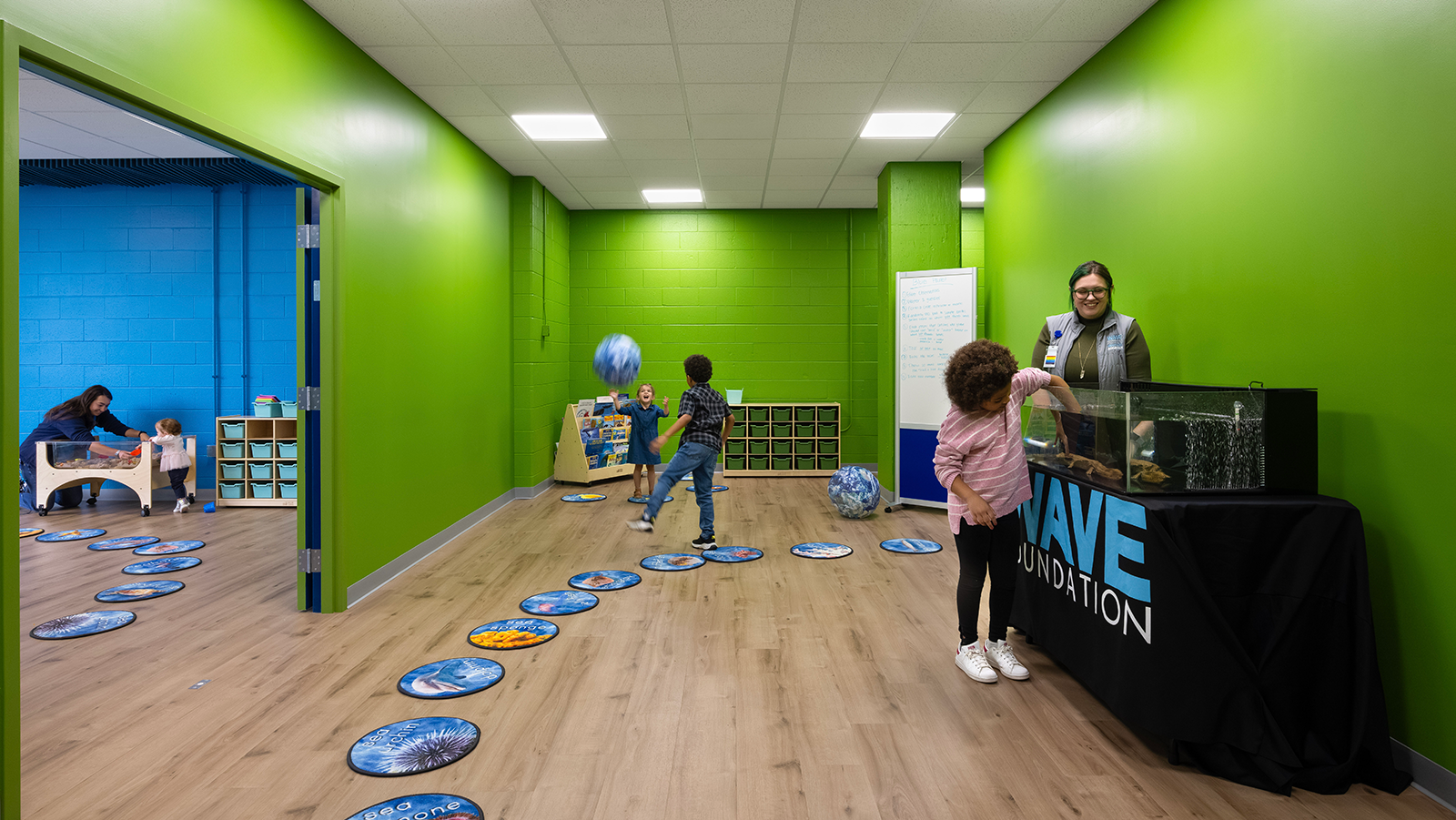 Photo showing context of how two classrooms connect. Children playing throughout and one is being supervised touching a marine animal in a tank.